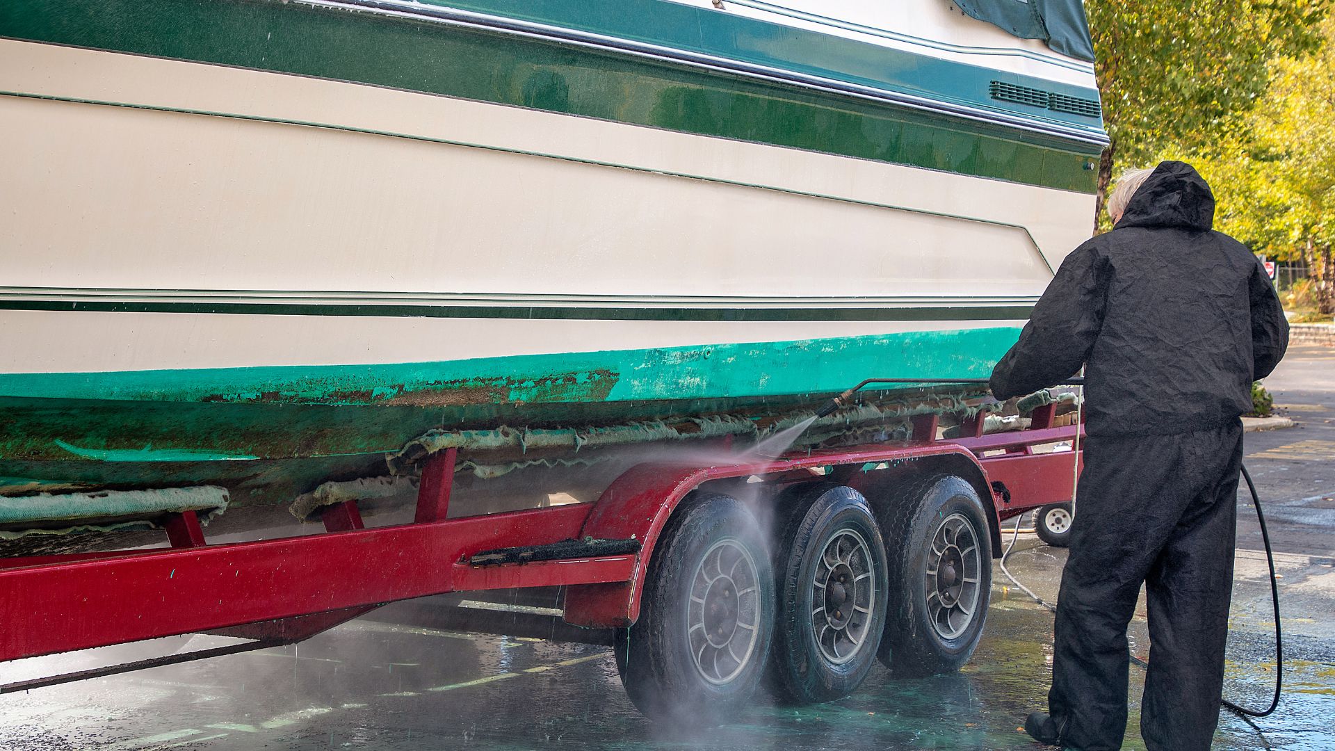 Person washing green and white boat trailer with high-pressure water hose