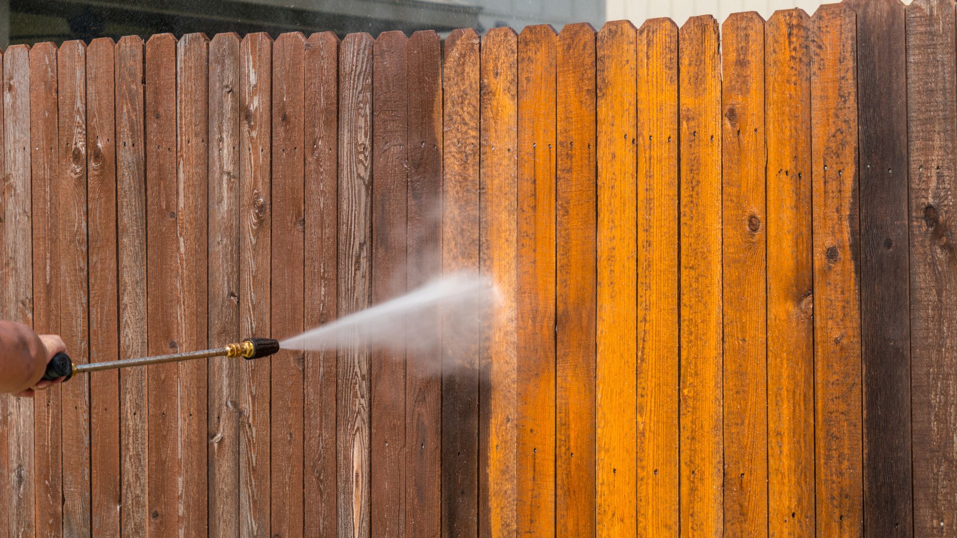 Pressure washer cleaning wooden fence, revealing bright orange wood underneath